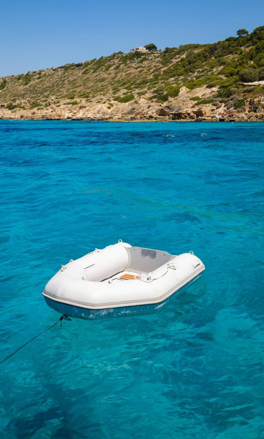 Small white inflatable boat floating on surface of clear turquoise sea near rocky sea against cloudless blue sky on sunny summer day